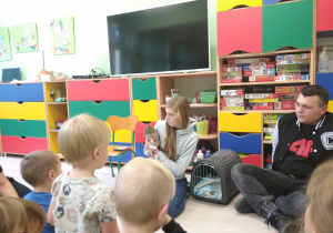 Dzieci siedzą na dywanie a Pani z Panem ze schroniska Cztery łapy opowiadają dzieciom o zwierzątkach.