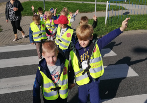 Dzieci przechodzą przez ulicę z podniesionymi rękoma, sygnalizując zbliżającym się pojazdom, że znajdują się na drodze