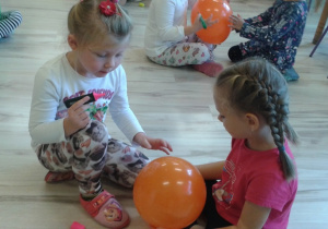 Dwie dziewczynki siedzą na podłodze. Jedna trzyma mazak w ręku a druga trzyma balon.