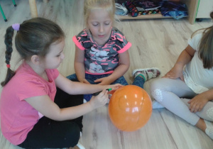 Trzy dziewczynki siedzą na podłodze jedna rysuje mazakiem po pomarańczowym balonie. Druga dziewczynka przygląda się jak jedna rysuje. Natomiast Trzecia z dziewczynek patrzy w drugą stronę.
