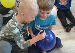 Jeden chłopiec rysuje mazakiem po niebieskim balonie a drugi trzyma balon.