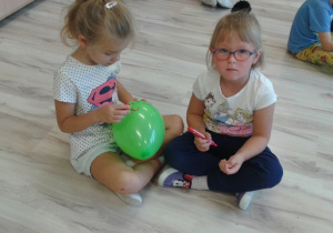 Jedna dziewczynka rysuje mazakiem po zielonym balonie a druga dziewczynka siedzi z mazakiem w ęku i czeka na swoją kolei.