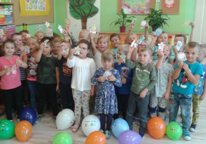 Dzieci z grupy VI pozują do zdjęcia trzymając wycięte odznaki w ręku. Na podłodze leżą balony.