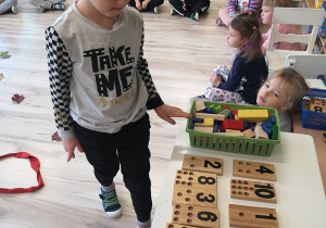 Chłopiec stoi przy stoliku na którym leżą podkładki z liczbami i odpowiednią liczba klocków.