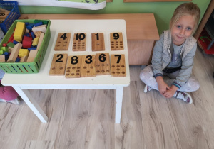 Uśmiechnięta dziewczynka siedzi na podłodze przy stoliku z klockami i liczbami.