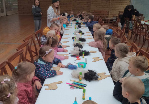 Dzieci na warsztatach z elementem sensoryki - pani tłumaczy dzieciom jak ozdobić drewnianą figurkę alpaki.