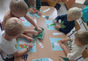 Dzieci wykonują pracę plastyczną przy stoliczkach w sali.