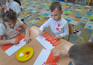 Dzieci wykonują pracę plastyczną, malują patyczkami kosmetycznymi na biało - czerwonych kartkach "Kwiaty dla Niepodległej"
