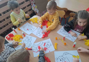 Dzieci przy stoliczkach wykonują prace plastyczne - uzupełniają wizerunek misia bibułą w kolorze żółtym i czerwonym