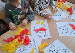 Dzieci przy stoliczkach wykonują prace plastyczne - uzupełniają wizerunek misia bibułą w kolorze żółtym i czerwonym