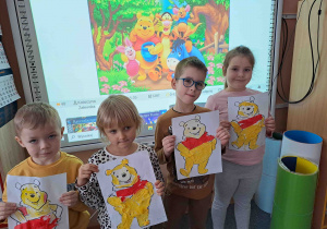 Dzieci prezentują wykonane prace na tle ilustracji z bajki o Puchatku