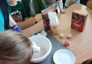 Na stoliku przygotowane są wszystkie składniki do wykonania pierniczków. Dzieci stoją wokoło stołu.