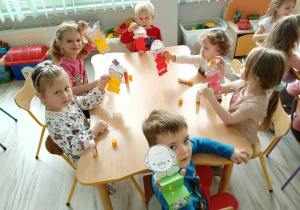 Dzieci siedzą przy stolikach i pokazują wykonane przez siebie prace.