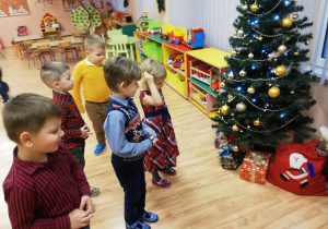 Dzieci stoją przed choinką i patrzą na leżące pod choinką prezenty