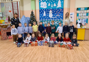 Grupa dzieci pozuje do zdjęcia wraz z nauczycielkami - dziewczynki siedzą na podłodze, trzymają w rękach prezenty, chłopcy stoją za dziewczynkami