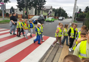 Dzieci ubrane w kamizelki odblaskowe przechodzą po pasach na drugą stronę ulicy.