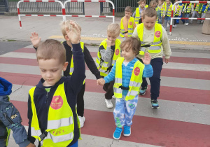 Dzieci ubrane w kamizelki odblaskowe przechodzą po pasach na drugą stronę ulicy.