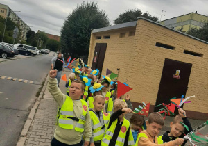 Dzieci idą po chodniku parami. Chłopcy machają chorągiewkami.