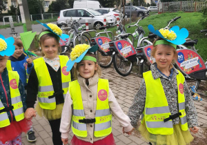 Dziewczynki idą w parach po chodniku. Na głowie mają opaski z dużym kwiatem niezapominajki.