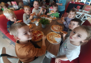 Dzieci siedzą przy stole i zajadają upieczone swoje pizzy.