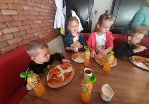 Dzieci siedzą przy stole i zajadają upieczone swoje pizzy.