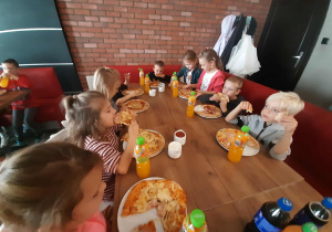 Dzieci siedzą przy stole i zajadają upieczone swoje pizzy.