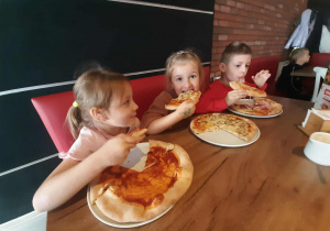Dzieci siedzą przy stole i zajadają upieczone swoje pizzy.