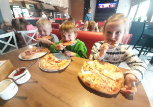 Dzieci siedzą przy stole i zajadają upieczone swoje pizzy.