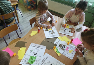 Czworo dzieci siedzi przy stoliku i każdy z nich wykleja kontur misia kolorowym papierem.