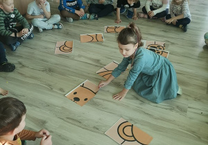 Dzieci siedzą w kole na podkładkach. Dziewczynka w środku koła układa z rozsypanki pluszowego misia.