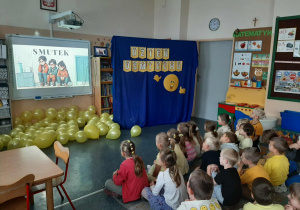 Dzieci siedzą na dywanie i oglądają na tablicy multimedialnej film edukacyjny o emocjach.