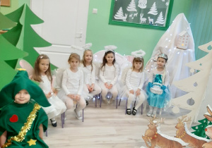 Po lewej stronie siedzi dziewczynka przebrana za choinkę, na wprost na krześle siedzą cztery aniołki, pani zima, wróżka