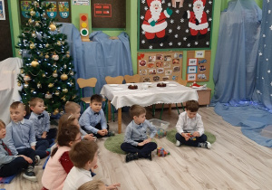 Na tle dekoracji świątecznej stoi na środku stolik przy nim na podłodze siedzi dwóch chłopców. Po lewej stronie siedzą pozostałe dzieci.