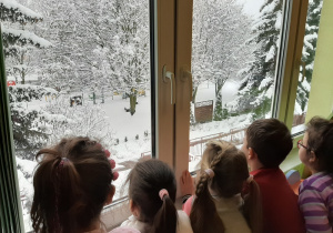 Pięcioro dzieci stoi przy oknie i podziwiają ogród przedszkolny w zimowej szacie.