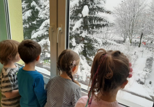 Czworo dzieci stoi przy oknie i podziwia krajobraz zimowy.