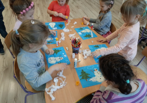 Sześcioro dzieci siedzi przy stoliku i wykonuje pracę plastyczną na temat zimowy krajobraz. Wyklejają kontur bałwana białą plasteliną oraz naklejają na chmurki białą bibułę.