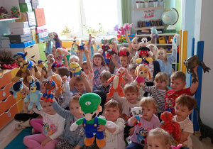 Dzieci siedzą na dywanie i podnoszą swoje maskotki.