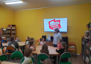 Dzieci siedzą przy stoliczkach i słuchają pani bibliotekarki