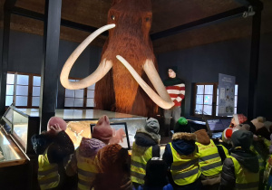 Dzieci w Muzeum Ziemi słuchają przewodnika, który opowiada ciekawostki
