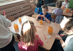 Dzieci wykonują lampiony przy stoliku. Malują farbami słoiki i naklejają ozdoby.