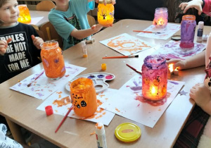 Dzieci wykonują lampiony przy stoliku. Malują farbami słoiki i naklejają ozdoby.