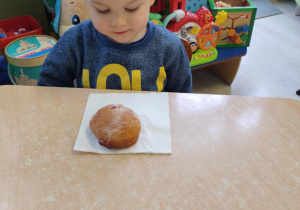 Chłopiec w niebieskiej koszulce siedzi przy stoliku, zjada pączka.