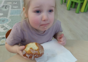 Dziewczynka w fioletowej koszulce je pączka przy stoliku.