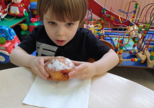 Chłopiec w granatowej koszulce je pączka przy stoliku.