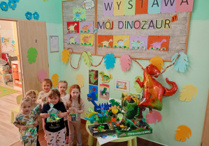 Dzieci z upominkami pozują do zdjęcia na tle wystawy plastycznej ,,Mój Dinozaur".