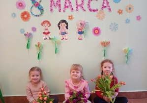 Dziewczynki pozują z kwiatami na tle dekoracji z okazji Dnia Kobiet.