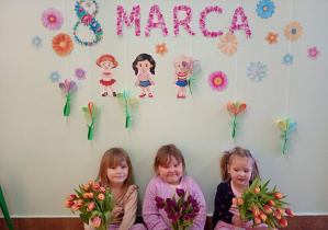 Dziewczynki pozują z kwiatami na tle dekoracji z okazji Dnia Kobiet.