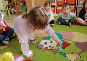 Dzieci na dywanie przyglądają się, jak ich koleżanka układa kreatywne materiały plastyczne na sylwecie twarzy dziewczynki.