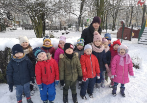 Dzieci wraz Panią wychowawczynią pozowały do zdjęcia. W tle widać zaśnieżony ogród przedszkolny.