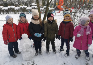 Sześcioro dzieci stoi uśmiechają się. Przed nimi stoi ulepiony bałwanek.